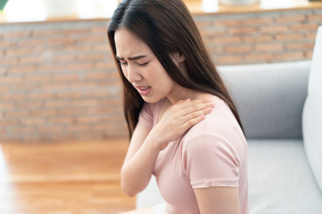 Young cute Asian girl having shoulder pain or ache problem sitting on sofa at home with painful.