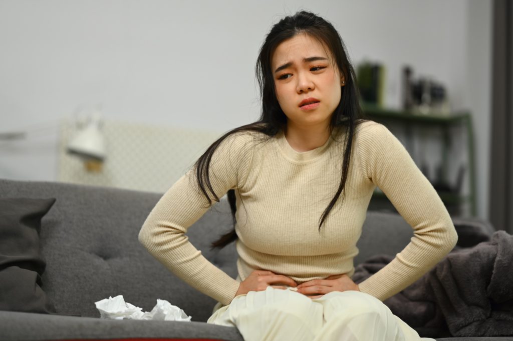 Mujer joven sentada en un sofá en casa sufriendo dolor de estómago, diarrea o problemas digestivos.